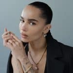 Channeling Zoë Kravitz vibes, a woman with slicked-back hair wears a black blazer, gold jewelry, and multiple rings. Tattoos on her hand add edge as she gazes calmly at the camera against a plain, light background.