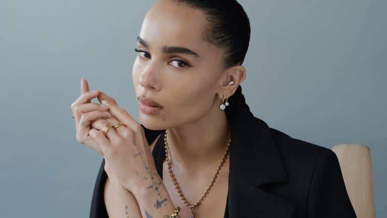 Channeling Zoë Kravitz vibes, a woman with slicked-back hair wears a black blazer, gold jewelry, and multiple rings. Tattoos on her hand add edge as she gazes calmly at the camera against a plain, light background.
