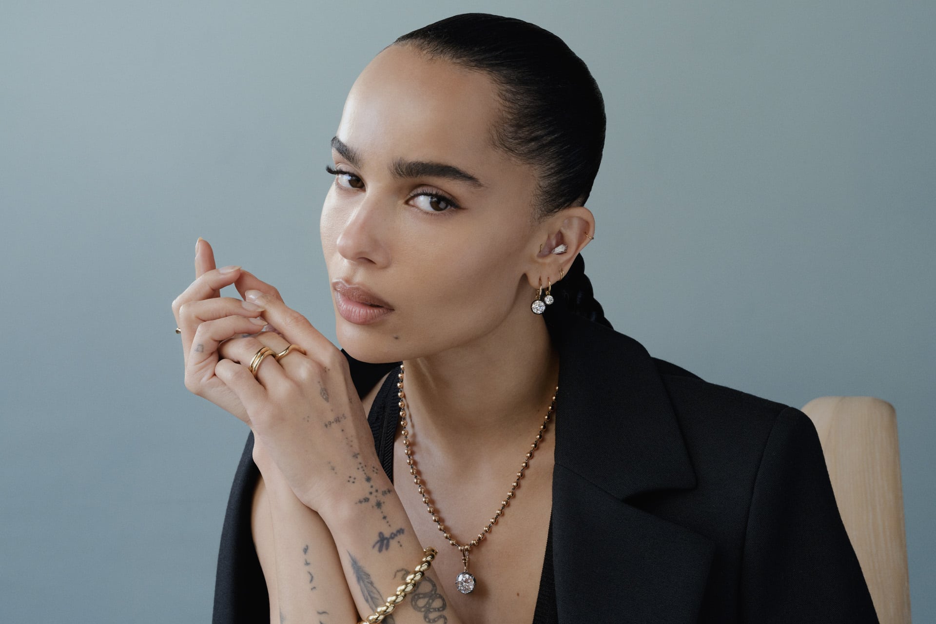 Channeling Zoë Kravitz vibes, a woman with slicked-back hair wears a black blazer, gold jewelry, and multiple rings. Tattoos on her hand add edge as she gazes calmly at the camera against a plain, light background.