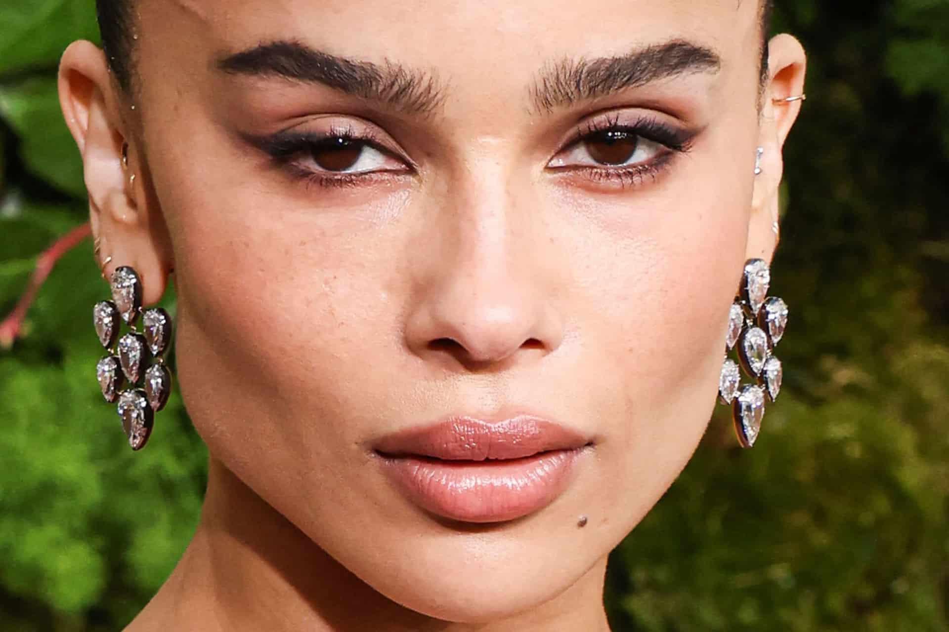 Close-up of a person with arched eyebrows, winged eyeliner, nude lipstick, and prominent earrings adorned with stones—evoking Zoë Kravitz’s chic style. The background is softly blurred with greenery.