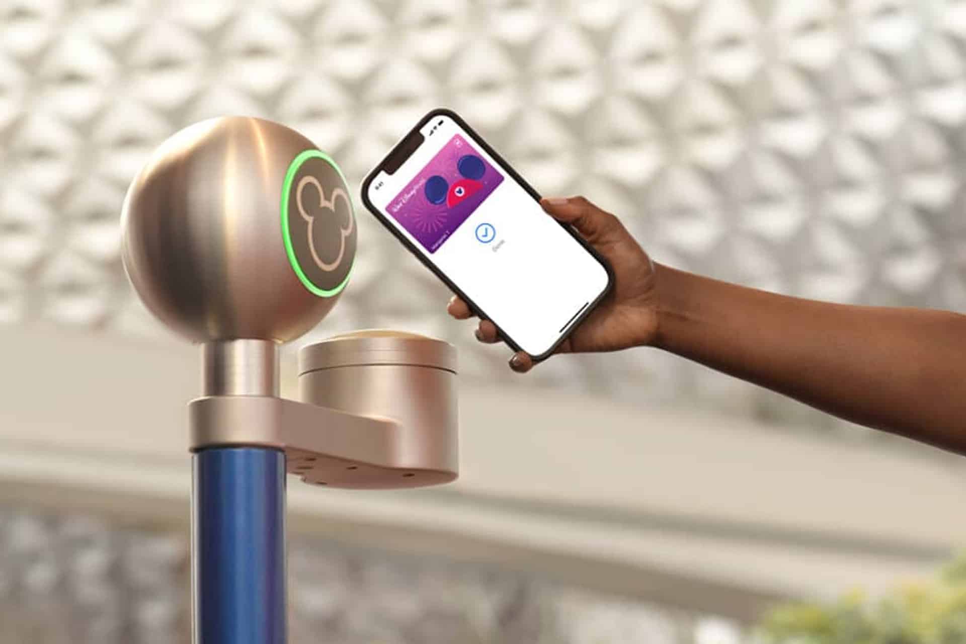 A person holds a smartphone with a Disney MagicMobile digital pass near a Mickey Mouse-shaped scanner, likely at a Disney World entrance. The background is blurred, emphasizing the seamless My Disney Experience scanning process.