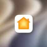 A white rounded square icon with an orange and yellow house symbol appears on a blurred background, representing Apple Home. The Apple logo is visible in the bottom right corner.