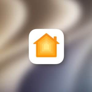A white rounded square icon with an orange and yellow house symbol appears on a blurred background, representing Apple Home. The Apple logo is visible in the bottom right corner.