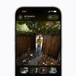 A smartphone screen displays HomeKit Secure Video camera footage of a person standing outside a wooden gate, surrounded by trees and leaves, with video clip thumbnails shown at the bottom.