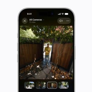 A smartphone screen displays HomeKit Secure Video camera footage of a person standing outside a wooden gate, surrounded by trees and leaves, with video clip thumbnails shown at the bottom.