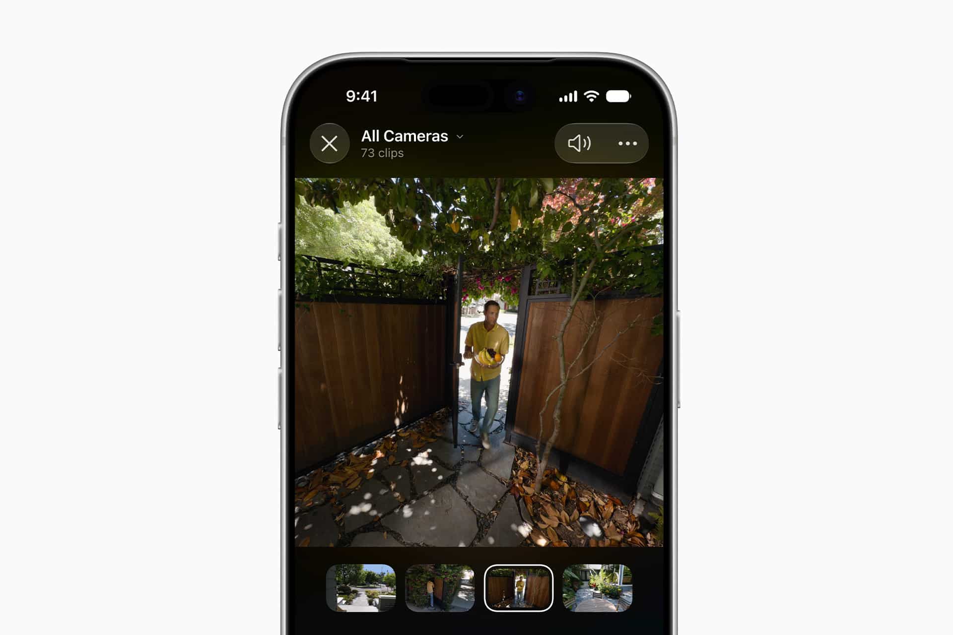 A smartphone screen displays HomeKit Secure Video camera footage of a person standing outside a wooden gate, surrounded by trees and leaves, with video clip thumbnails shown at the bottom.