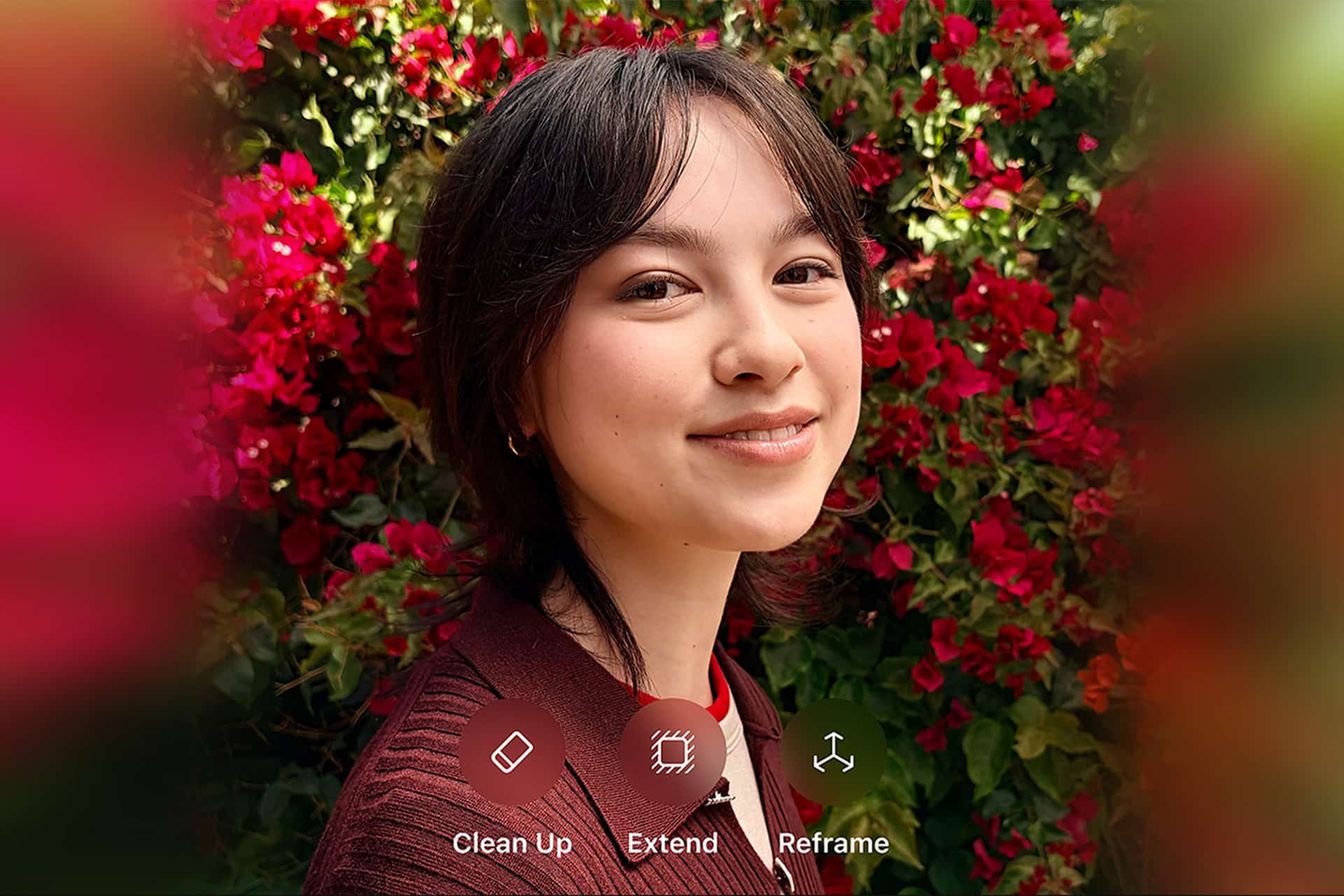 A young person with short dark hair smiles in front of a lush background of bright red flowers. On-screen iOS 27 AI photo editing options labeled Clean Up, Extend, and Reframe appear at the bottom of the image.
