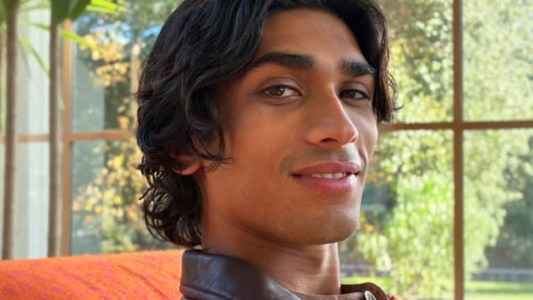 A person with medium-length dark hair and a slight smile sits indoors near a large window. Sunlight brightens green trees and leafy plants in the background. The person wears a brown-collared shirt, captured using iOS 27 AI photo editing in the Photos app.