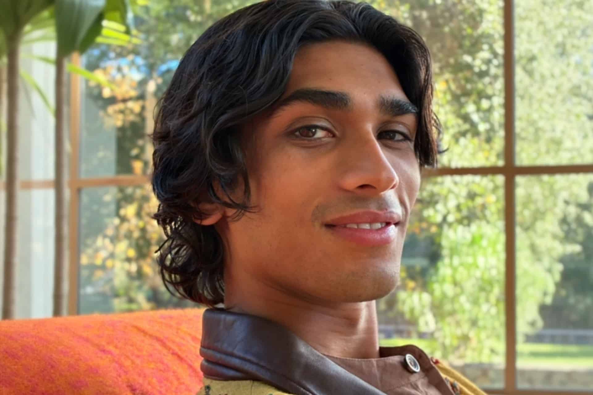 A person with medium-length dark hair and a slight smile sits indoors near a large window. Sunlight brightens green trees and leafy plants in the background. The person wears a brown-collared shirt, captured using iOS 27 AI photo editing in the Photos app.