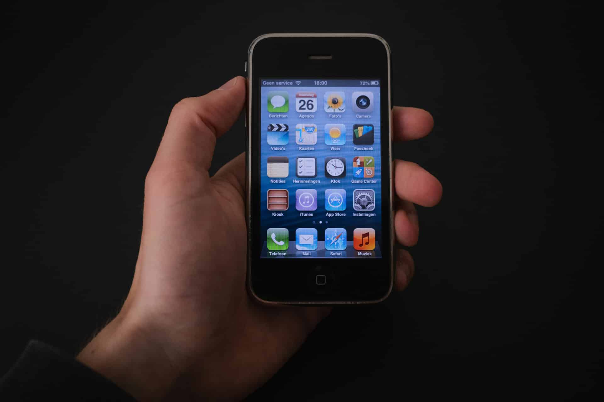 A hand holds an older model black iPhone with a home button, displaying a home screen with various app icons against a dark background—a testament to the design vision of Steve Jobs and Apple leadership.