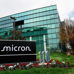 A large glass office building with a “Micron” sign in front, surrounded by green grass, red and white flowers, and trees with autumn leaves on a cloudy day—home to innovations in DRAM and NAND flash memory.