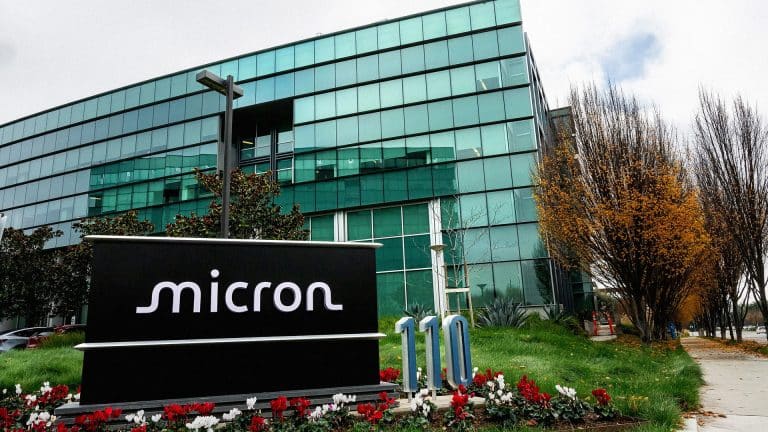 A large glass office building with a “Micron” sign in front, surrounded by green grass, red and white flowers, and trees with autumn leaves on a cloudy day—home to innovations in DRAM and NAND flash memory.