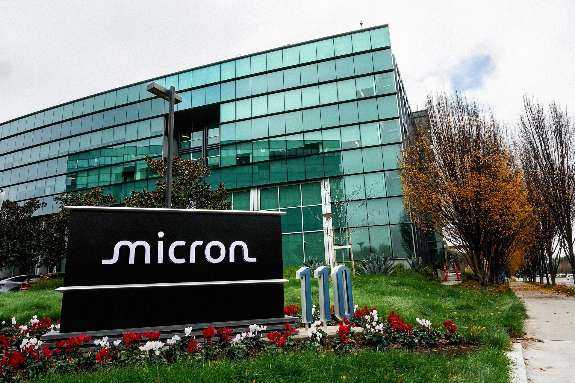 A large glass office building with a “Micron” sign in front, surrounded by green grass, red and white flowers, and trees with autumn leaves on a cloudy day—home to innovations in DRAM and NAND flash memory.