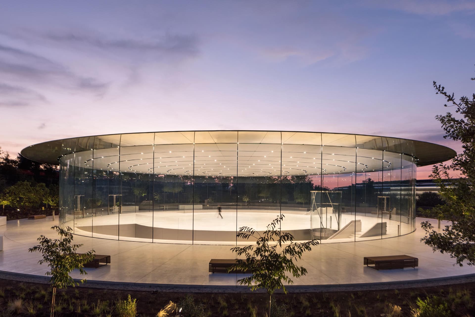 A circular glass building with a flat roof, minimalistic design, and empty interior, reminiscent of the Steve Jobs Theater, is surrounded by greenery at sunset.