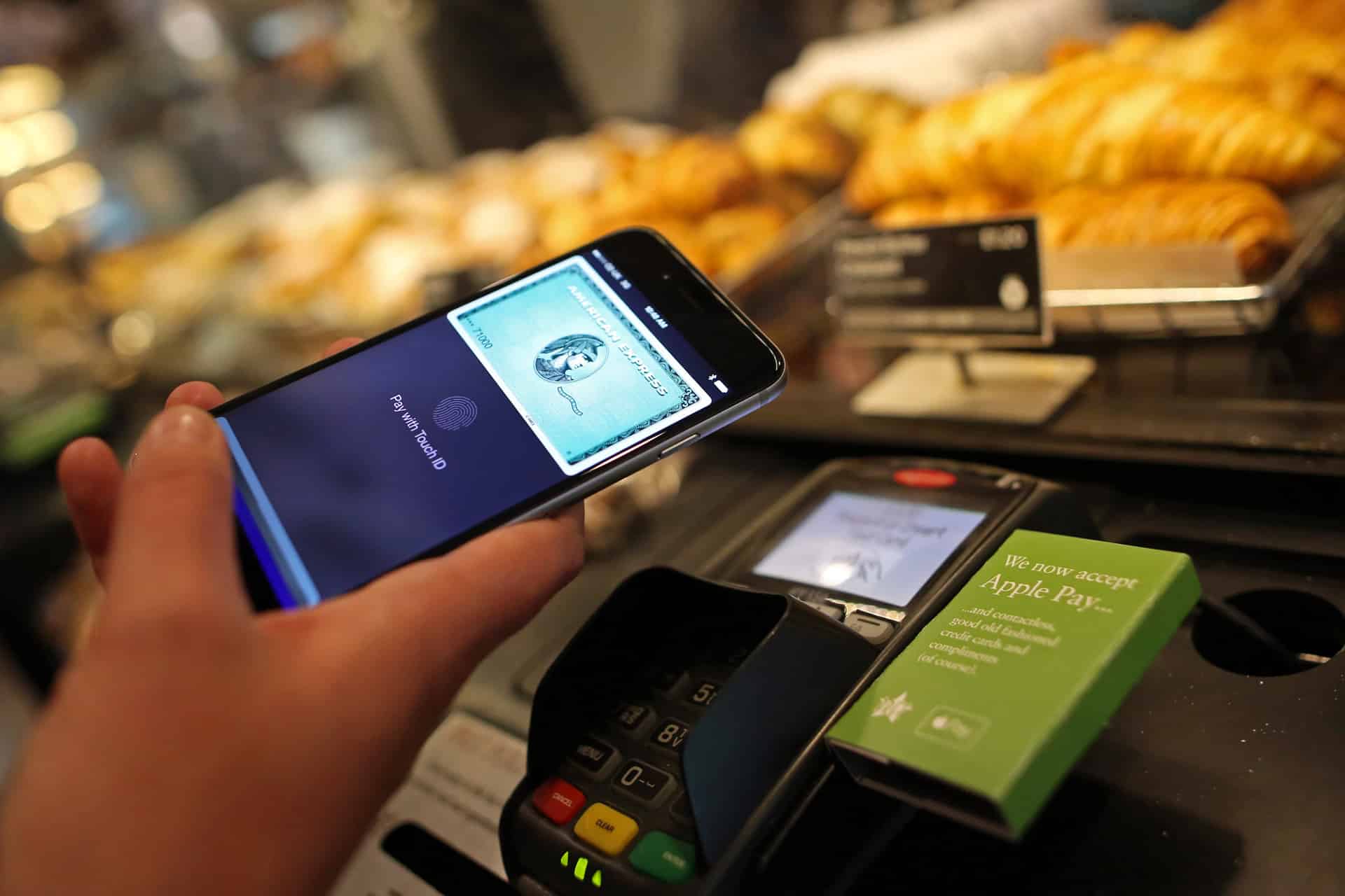A person holds a smartphone with Apple Pay open over a payment terminal at a bakery counter, preparing an American Express contactless payment. Pastries are visible in the background.
