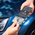 A person holds an American Express credit card near a smartphone displaying a $75.00 payment screen in Apple Wallet, with a blue car in the background, suggesting a contactless payment transaction.