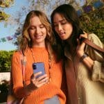 Two women stand outdoors, smiling as they look at a smartphone together. With bags over their shoulders and colorful banners behind them, they seem excited, possibly exploring new favorites through App Store discovery in a festive setting.