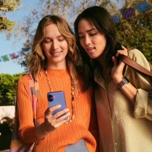 Two women stand outdoors, smiling as they look at a smartphone together. With bags over their shoulders and colorful banners behind them, they seem excited, possibly exploring new favorites through App Store discovery in a festive setting.