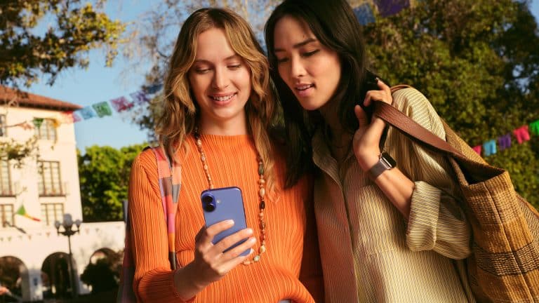 Two women stand outdoors, smiling as they look at a smartphone together. With bags over their shoulders and colorful banners behind them, they seem excited, possibly exploring new favorites through App Store discovery in a festive setting.
