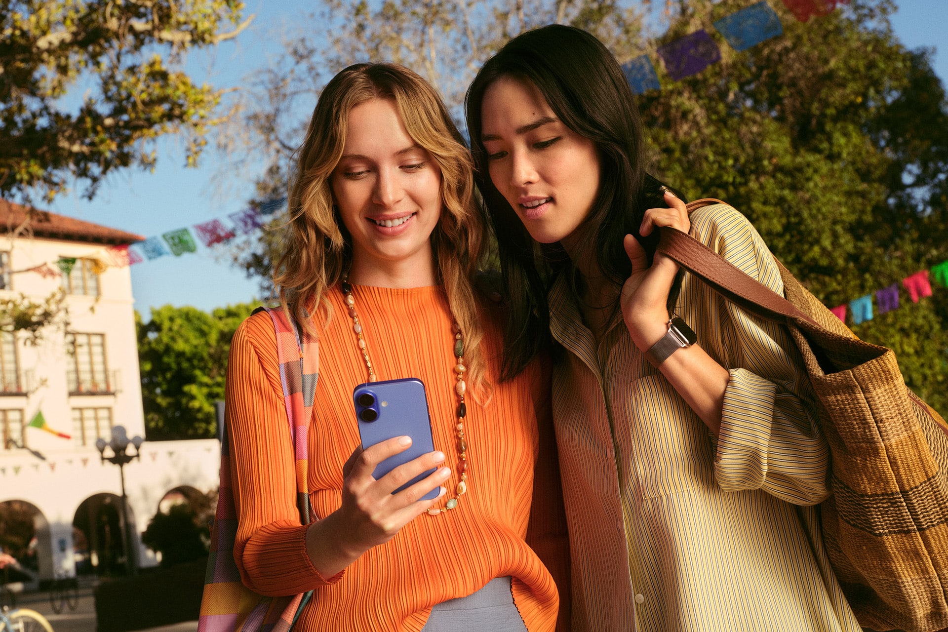 Two women stand outdoors, smiling as they look at a smartphone together. With bags over their shoulders and colorful banners behind them, they seem excited, possibly exploring new favorites through App Store discovery in a festive setting.