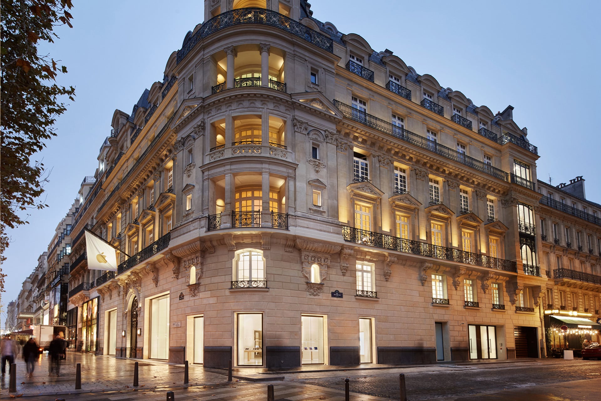 Elegant, historic Parisian building on a street corner at dusk, with warm lights glowing from large windows and ornate balconies—blending classic charm with Apple Store architecture. Pedestrians stroll nearby, passing a white flag with a gold emblem by the entrance.