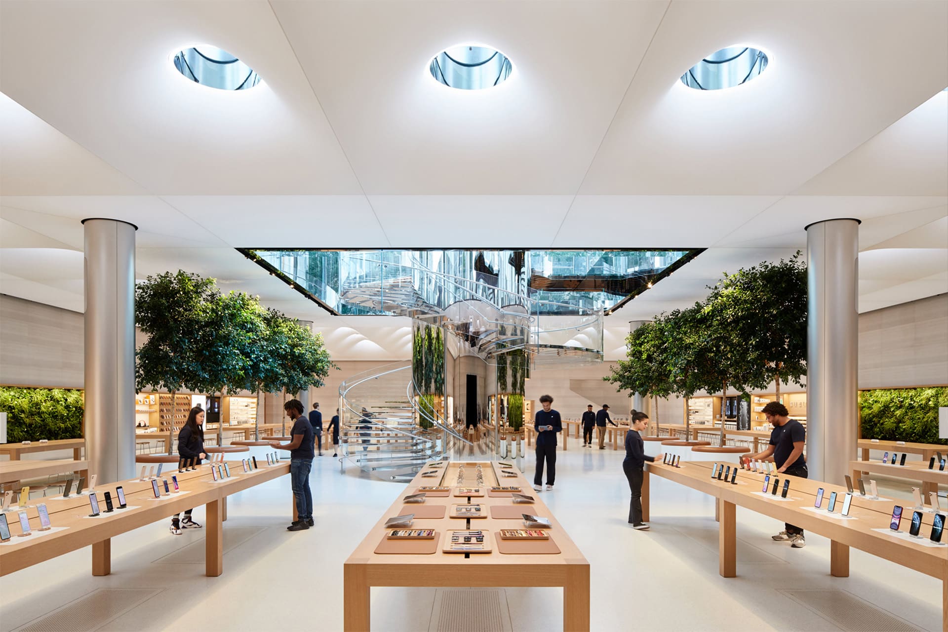 A modern Apple retail store with bright lighting, large tables displaying electronic devices, several staff members assisting customers, tall indoor trees, and a glass spiral staircase in the center—a reflection of Steve Jobs’ visionary design influence.