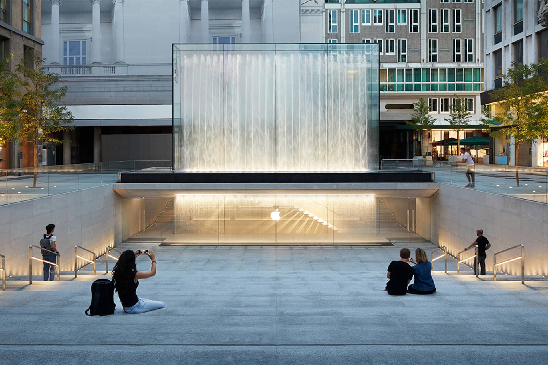 People sit on wide steps facing a modern glass cube, a striking example of Apple Store architecture. Below, the entrance to an Apple Store glows through glass walls. Urban buildings and trees complete this iconic Apple retail scene.