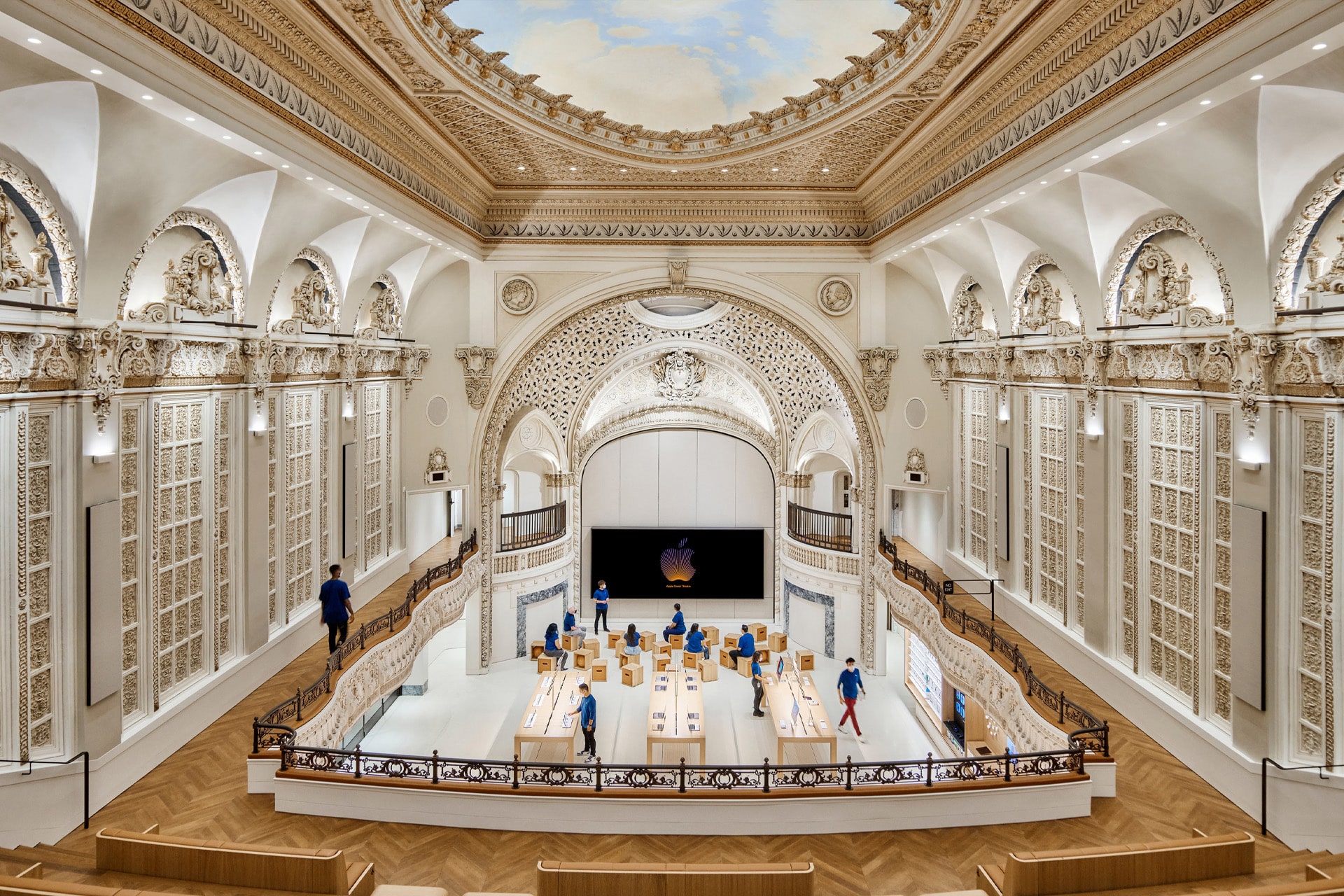 A grand, ornate hall with intricate architectural details and a painted ceiling now showcases stunning Apple Store architecture, featuring wooden tables, products on display, and several staff members in blue shirts.