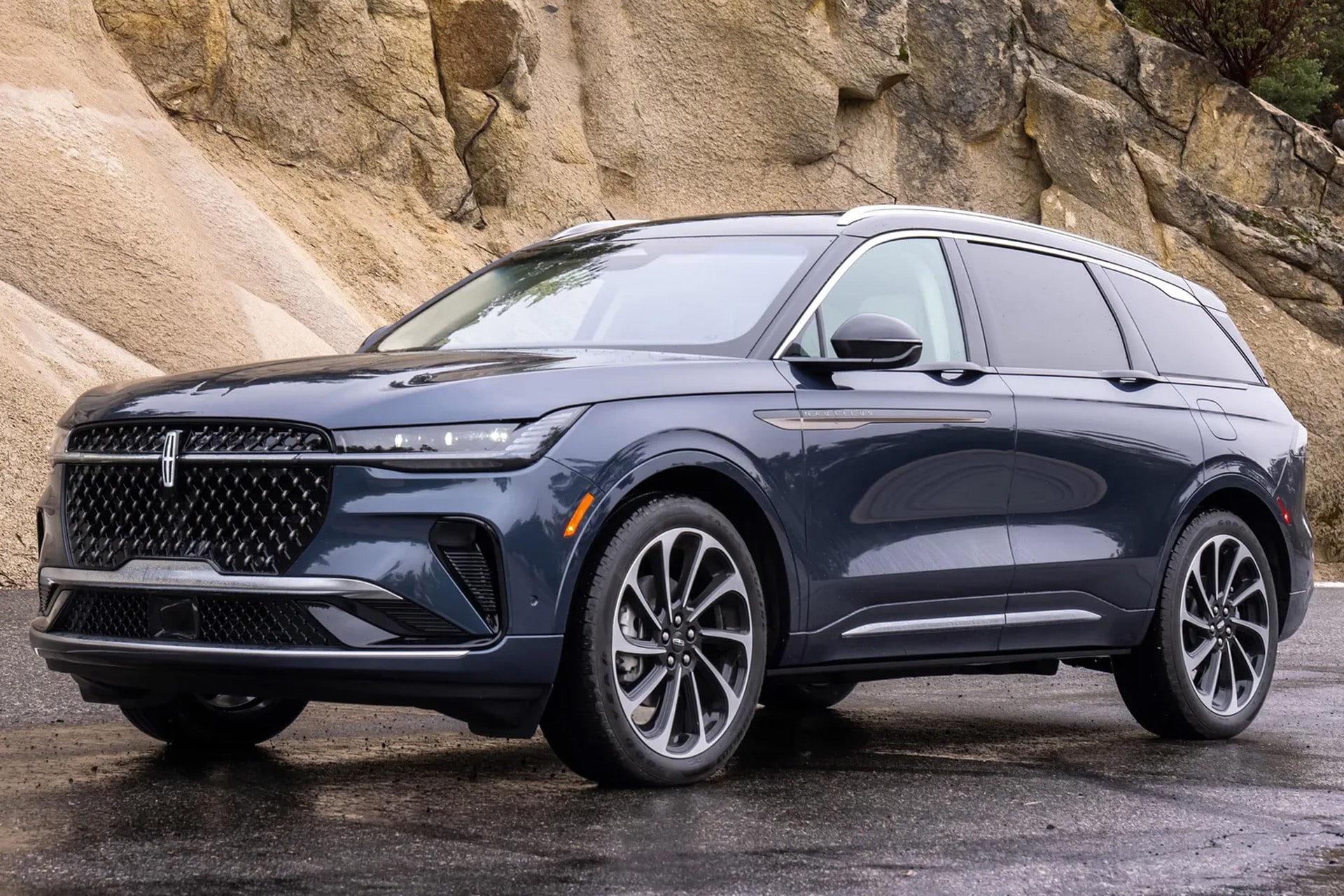 A dark blue SUV with chrome accents is parked on a wet road in front of a rocky hillside. The vehicle features a large front grille, sleek, modern design, and seamless Apple Maps integration for enhanced navigation on every journey.