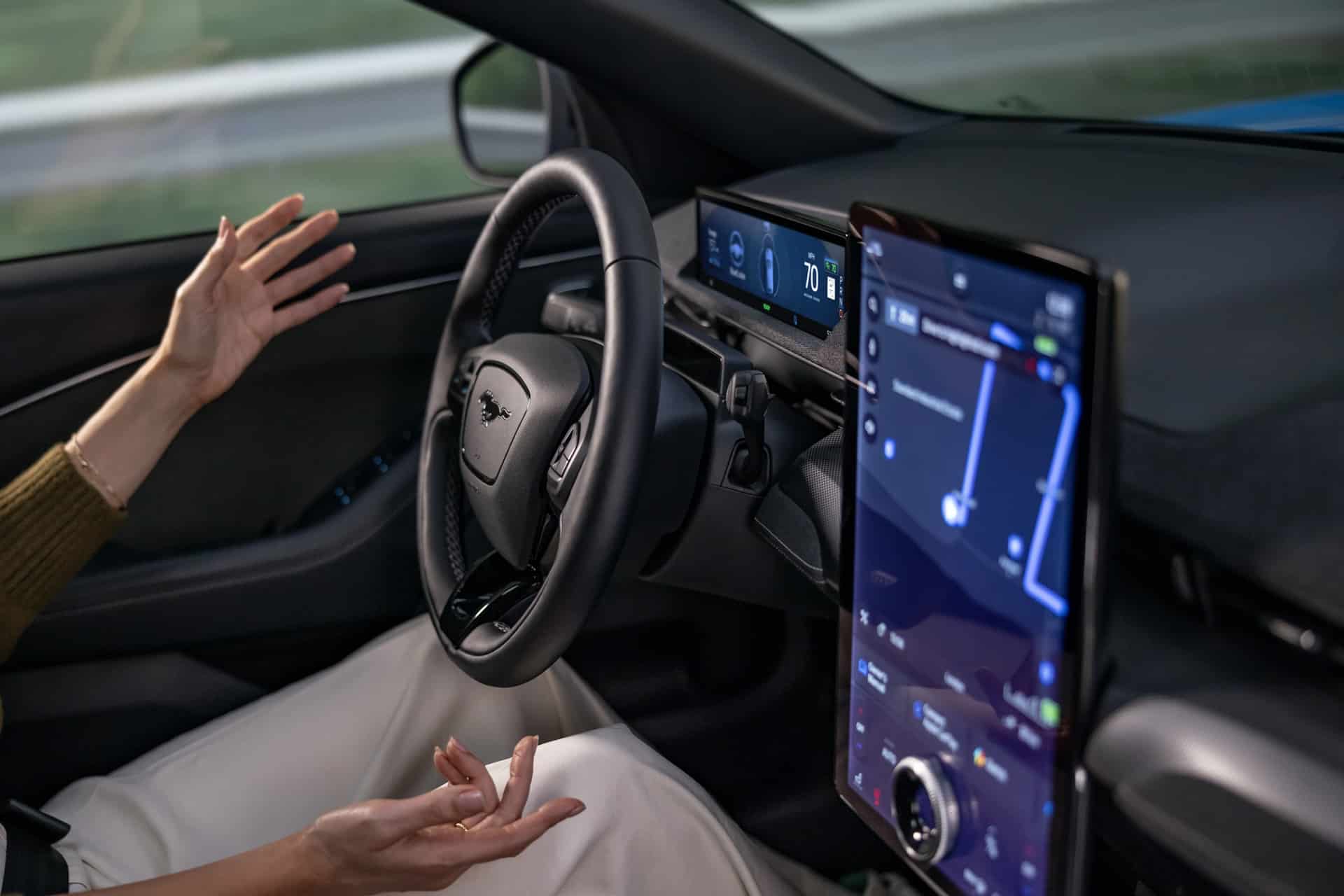 A person sits in the driver's seat of a car with their hands off the wheel, indicating autonomous driving. The dashboard displays speed and navigation on a large touchscreen, featuring Ford Apple Maps integration for seamless route guidance.