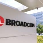 A sign with the Broadcom logo and name is displayed outside an office building, highlighting the site’s importance amid news of the Broadcom deal for Apple chips, with trees, cars, and another modern building in the background.
