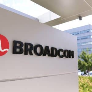 A sign with the Broadcom logo and name is displayed outside an office building, highlighting the site’s importance amid news of the Broadcom deal for Apple chips, with trees, cars, and another modern building in the background.