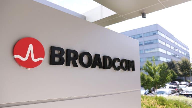 A sign with the Broadcom logo and name is displayed outside an office building, highlighting the site’s importance amid news of the Broadcom deal for Apple chips, with trees, cars, and another modern building in the background.