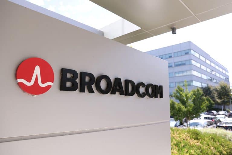 A sign with the Broadcom logo and name is displayed outside an office building, highlighting the site’s importance amid news of the Broadcom deal for Apple chips, with trees, cars, and another modern building in the background.