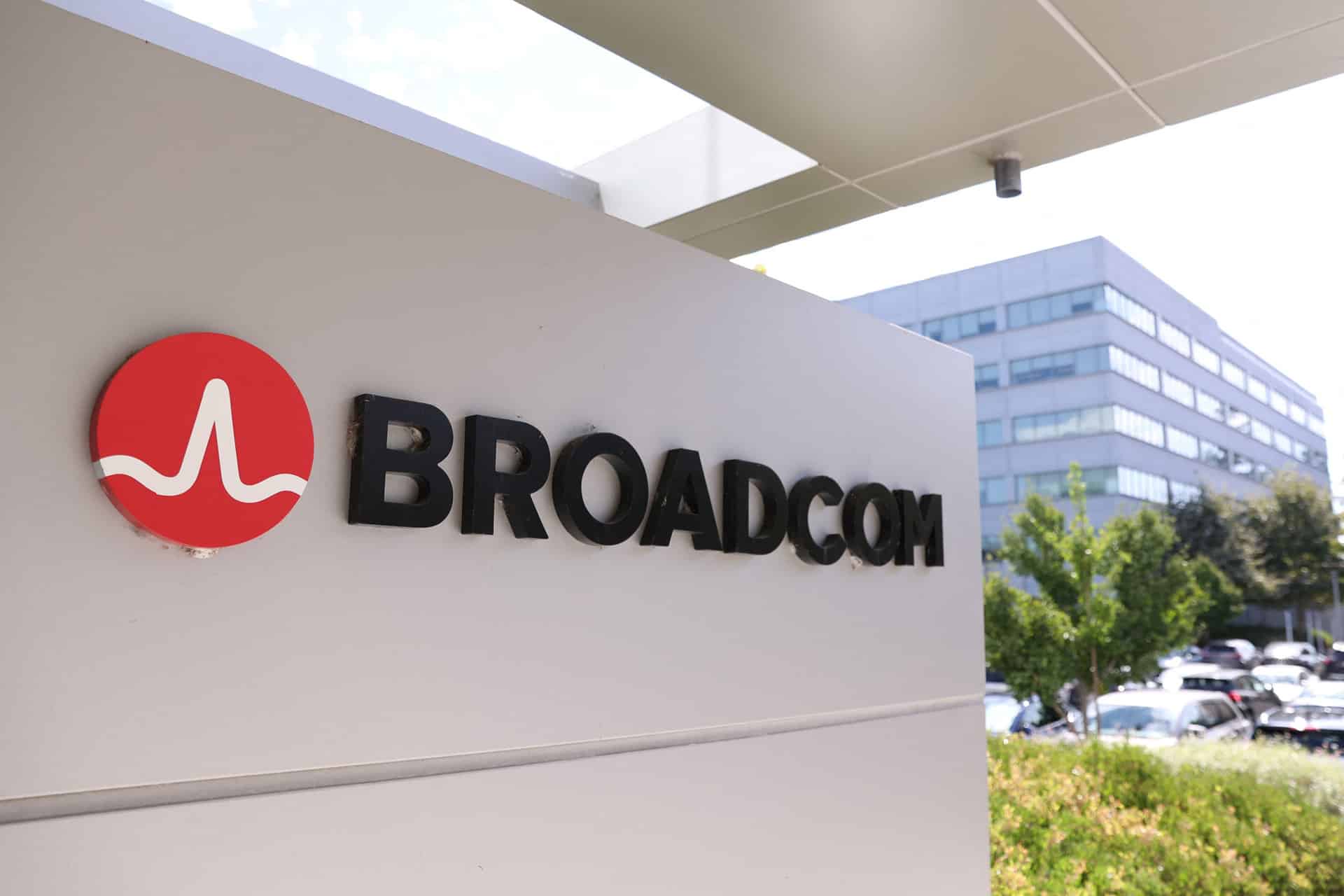 A sign with the Broadcom logo and name is displayed outside an office building, highlighting the site’s importance amid news of the Broadcom deal for Apple chips, with trees, cars, and another modern building in the background.
