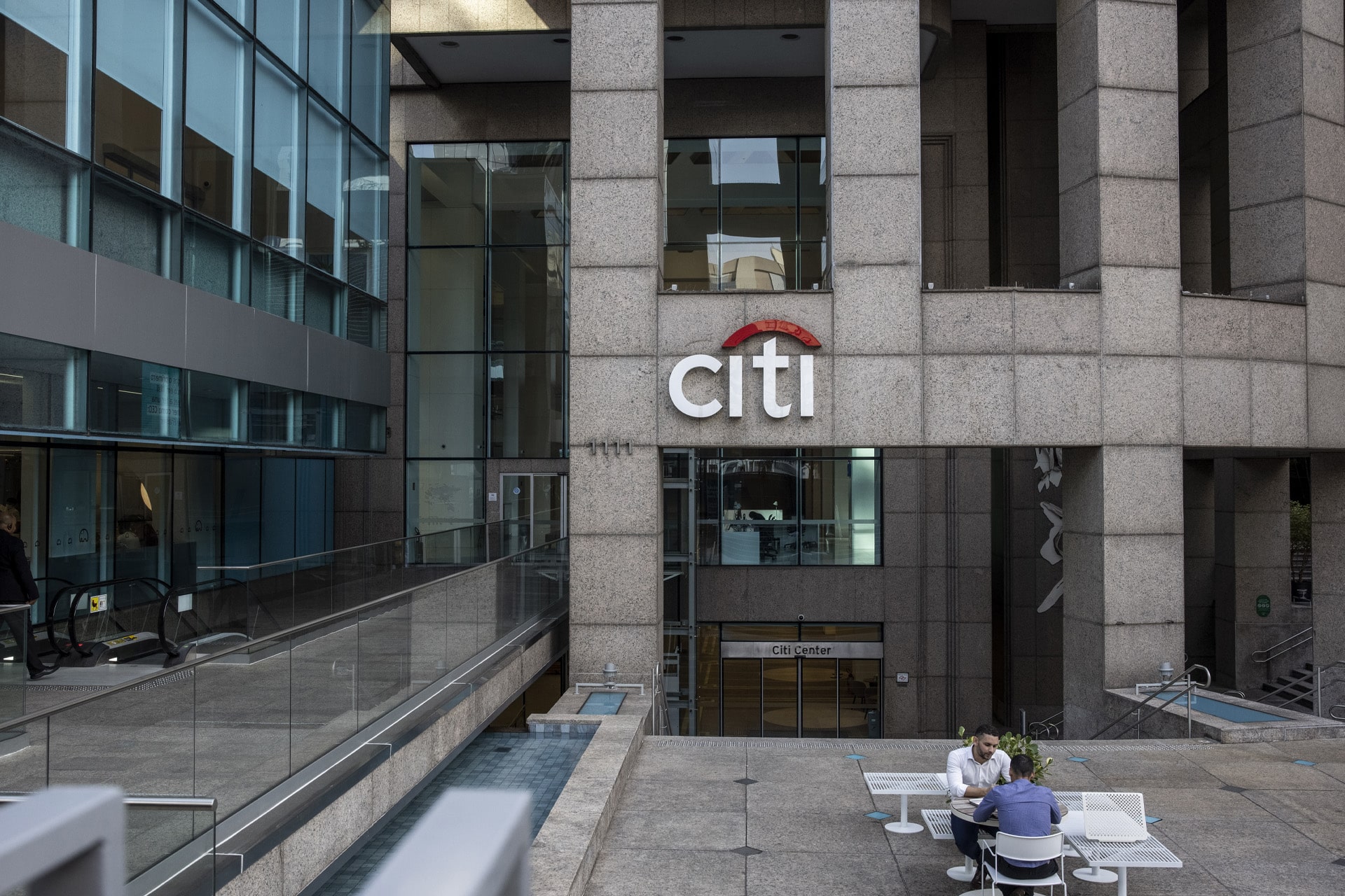 Citi Apple target - Two people sit at a white outdoor table in front of a large modern building with glass windows and a prominent "Citi" sign, engaged in conversation about technology trends like Siri AI and the latest movements in Apple stock (AAPL).