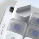 Close-up of a metallic knob and translucent buttons on a sleek OpenAI device. Text on the device reads, “OpenAI | 2026.” The design is modern, with a soft, neutral color palette and clear, blue-accented buttons—echoing the refined sensibilities often associated with Jony Ive’s hardware innovations.