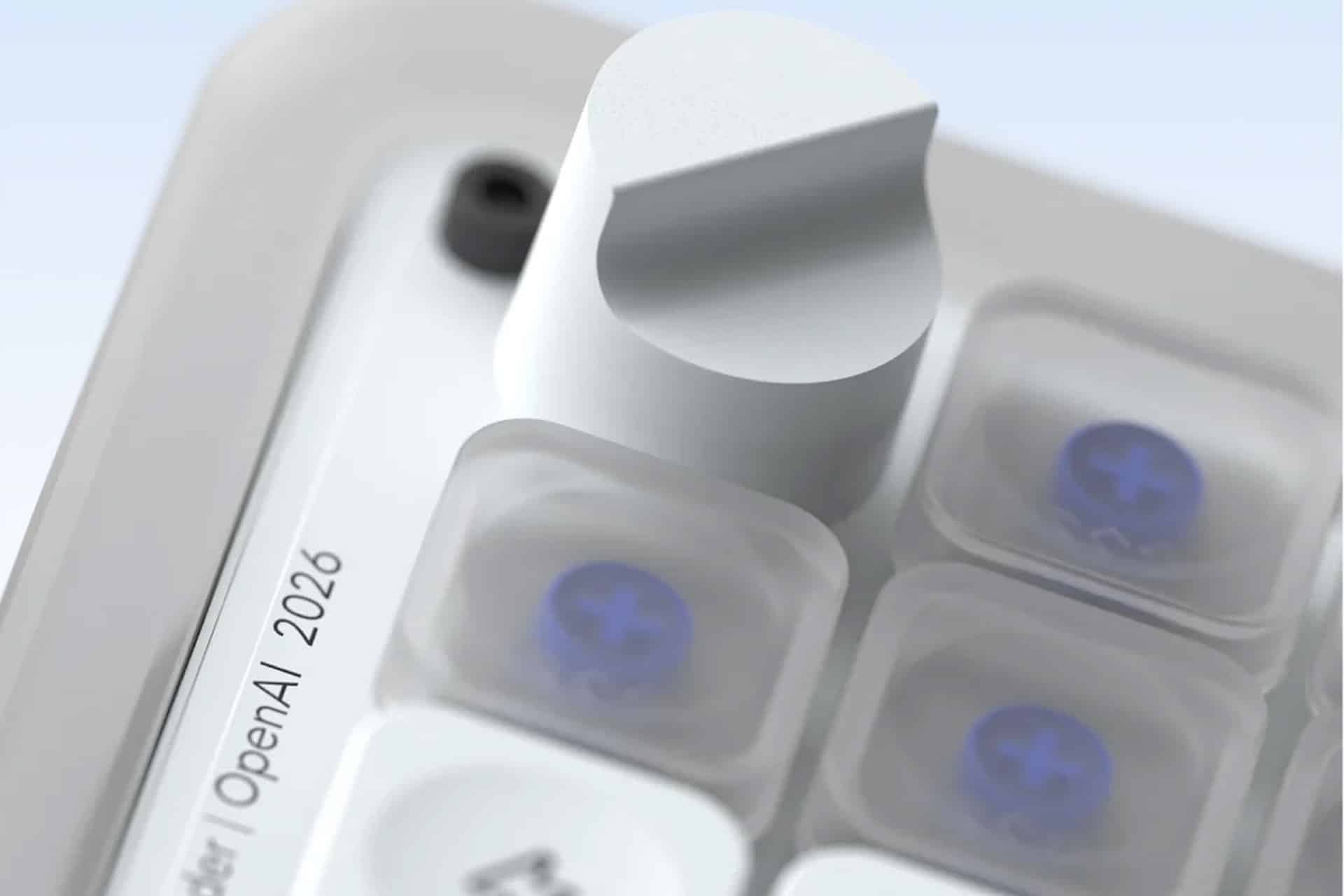 Close-up of a metallic knob and translucent buttons on a sleek OpenAI device. Text on the device reads, “OpenAI | 2026.” The design is modern, with a soft, neutral color palette and clear, blue-accented buttons—echoing the refined sensibilities often associated with Jony Ive’s hardware innovations.