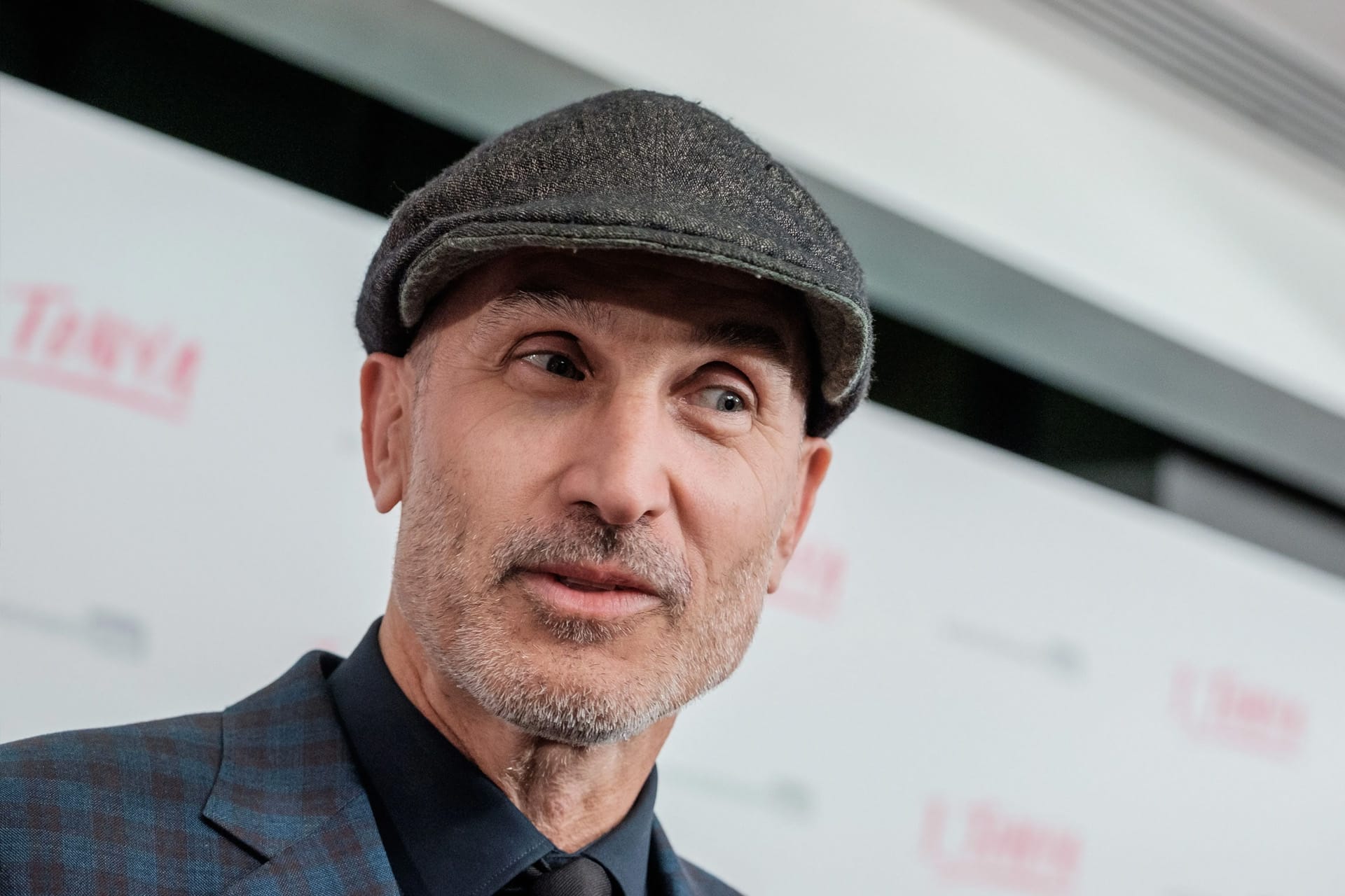 A man with a trimmed beard wears a flat cap, dark shirt, and checked blazer, standing in front of a white background with red and gray text—reminiscent of a scene from Apple TV thriller Guilty Creatures starring Julia Garner.