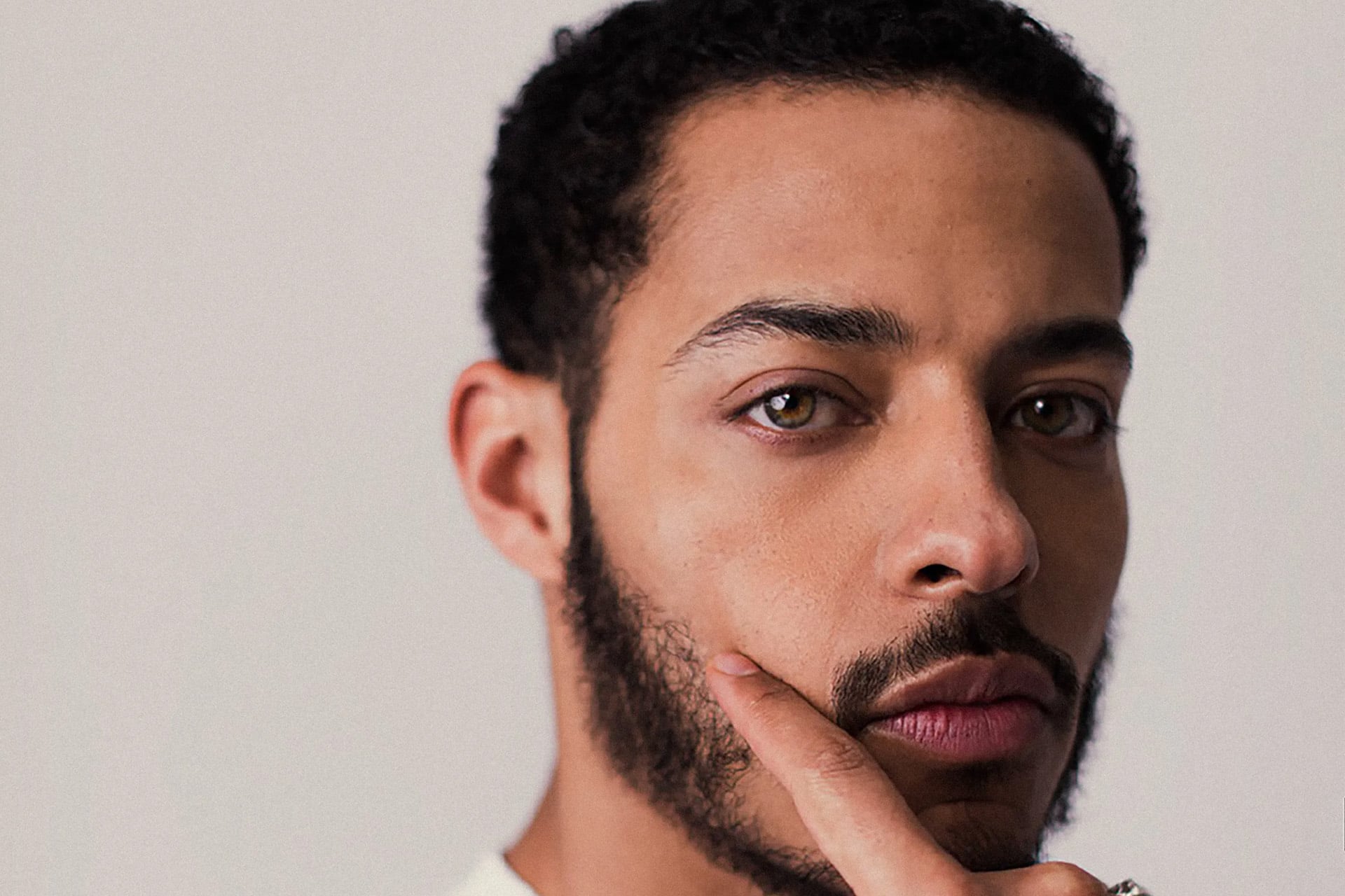 A close-up of a man with short curly hair and a beard, touching his face with one finger while looking thoughtfully toward the camera—reminiscent of an Apple TV thriller character created by Alex Cary.
