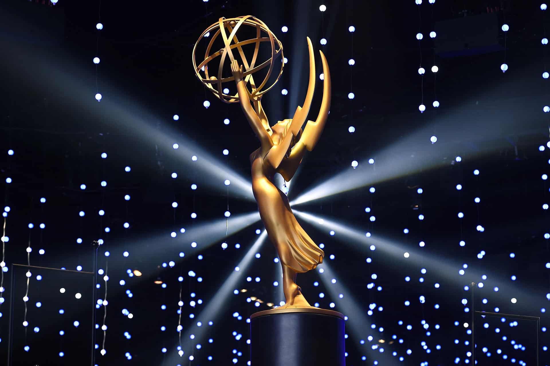 A gold Emmy Award statue is displayed on a pedestal, with dramatic lighting and numerous small, glowing lights in the background, creating a star-like effect reminiscent of prestigious Apple TV awards.