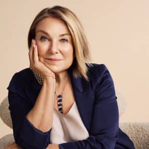 A woman with blonde hair in a navy blazer rests her head on her hand, smiling softly, sitting on a light-colored chair against a beige background. She wears a necklace and a light blouse, exuding the thoughtful charm reminiscent of Esther Perel from an Apple TV dating series.