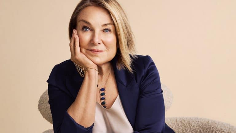 A woman with blonde hair in a navy blazer rests her head on her hand, smiling softly, sitting on a light-colored chair against a beige background. She wears a necklace and a light blouse, exuding the thoughtful charm reminiscent of Esther Perel from an Apple TV dating series.