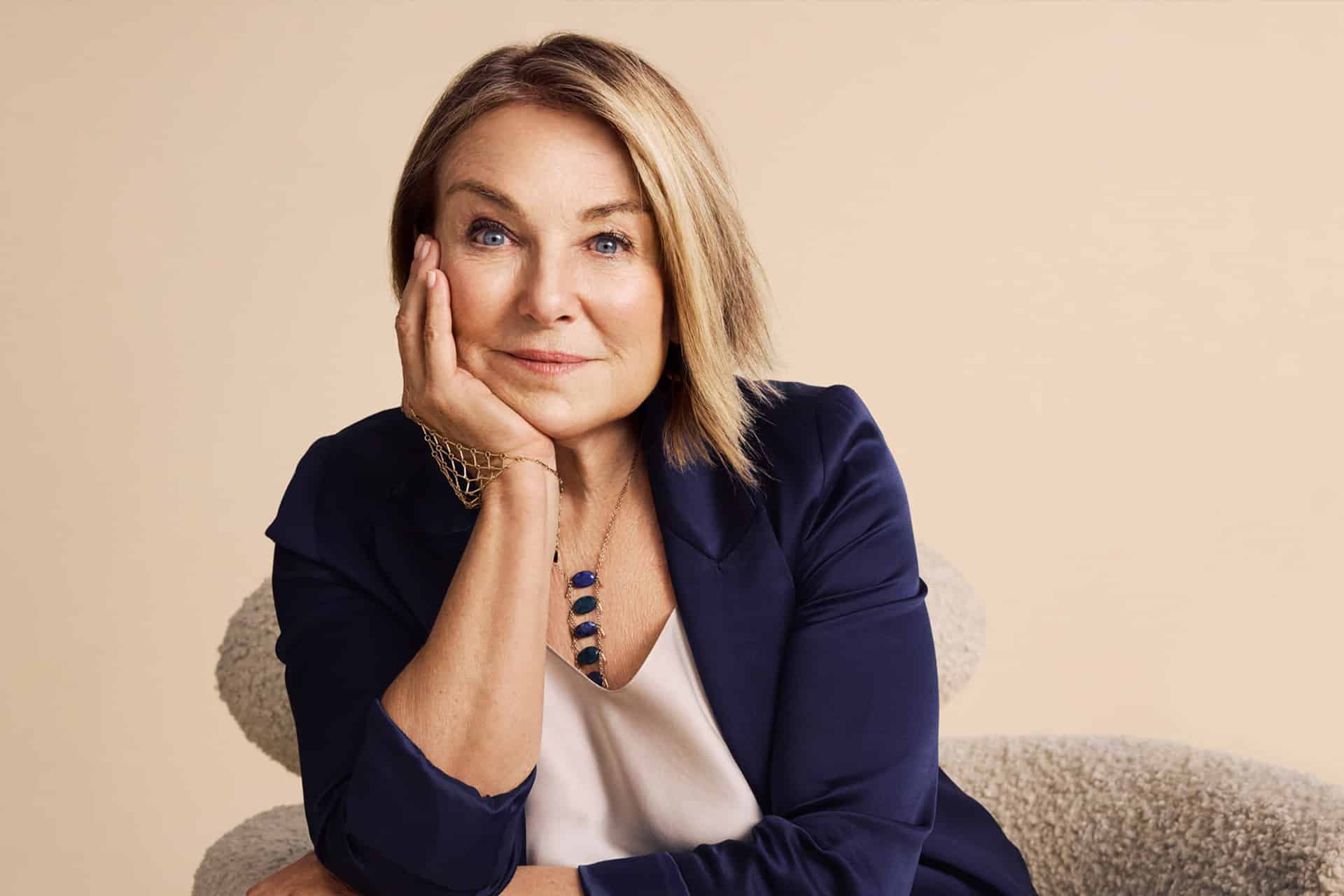 A woman with blonde hair in a navy blazer rests her head on her hand, smiling softly, sitting on a light-colored chair against a beige background. She wears a necklace and a light blouse, exuding the thoughtful charm reminiscent of Esther Perel from an Apple TV dating series.