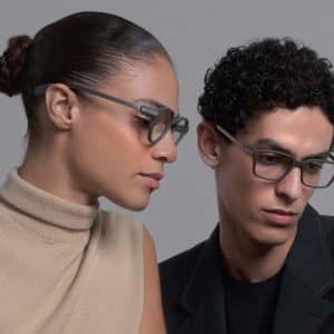 A woman in a beige sleeveless top and a man in a black blazer, both wearing smart glasses, look at a laptop screen together. The woman’s hair is in a bun and the man has curly hair.