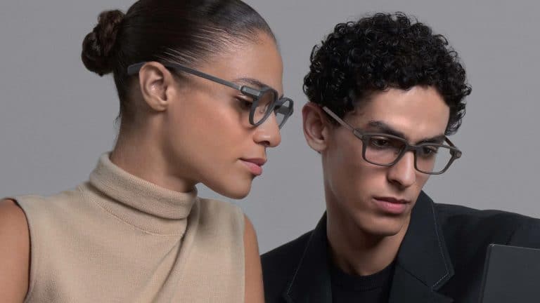 A woman in a beige sleeveless top and a man in a black blazer, both wearing smart glasses, look at a laptop screen together. The woman’s hair is in a bun and the man has curly hair.