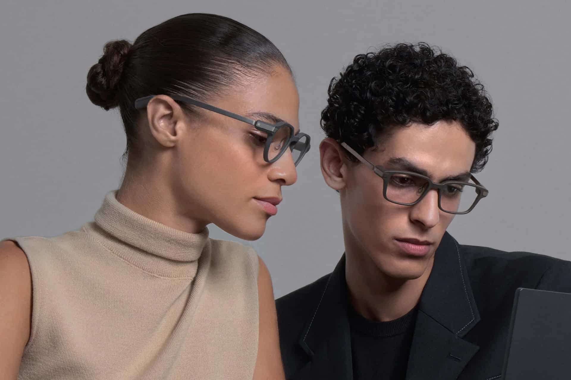 A woman in a beige sleeveless top and a man in a black blazer, both wearing smart glasses, look at a laptop screen together. The woman’s hair is in a bun and the man has curly hair.