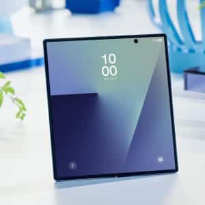 A foldable smartphone featuring Samsung creaseless displays stands open on a white surface, displaying its home screen with the time 10:00. The background includes a plant, a blue vase, and books.