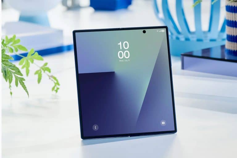 A foldable smartphone featuring Samsung creaseless displays stands open on a white surface, displaying its home screen with the time 10:00. The background includes a plant, a blue vase, and books.