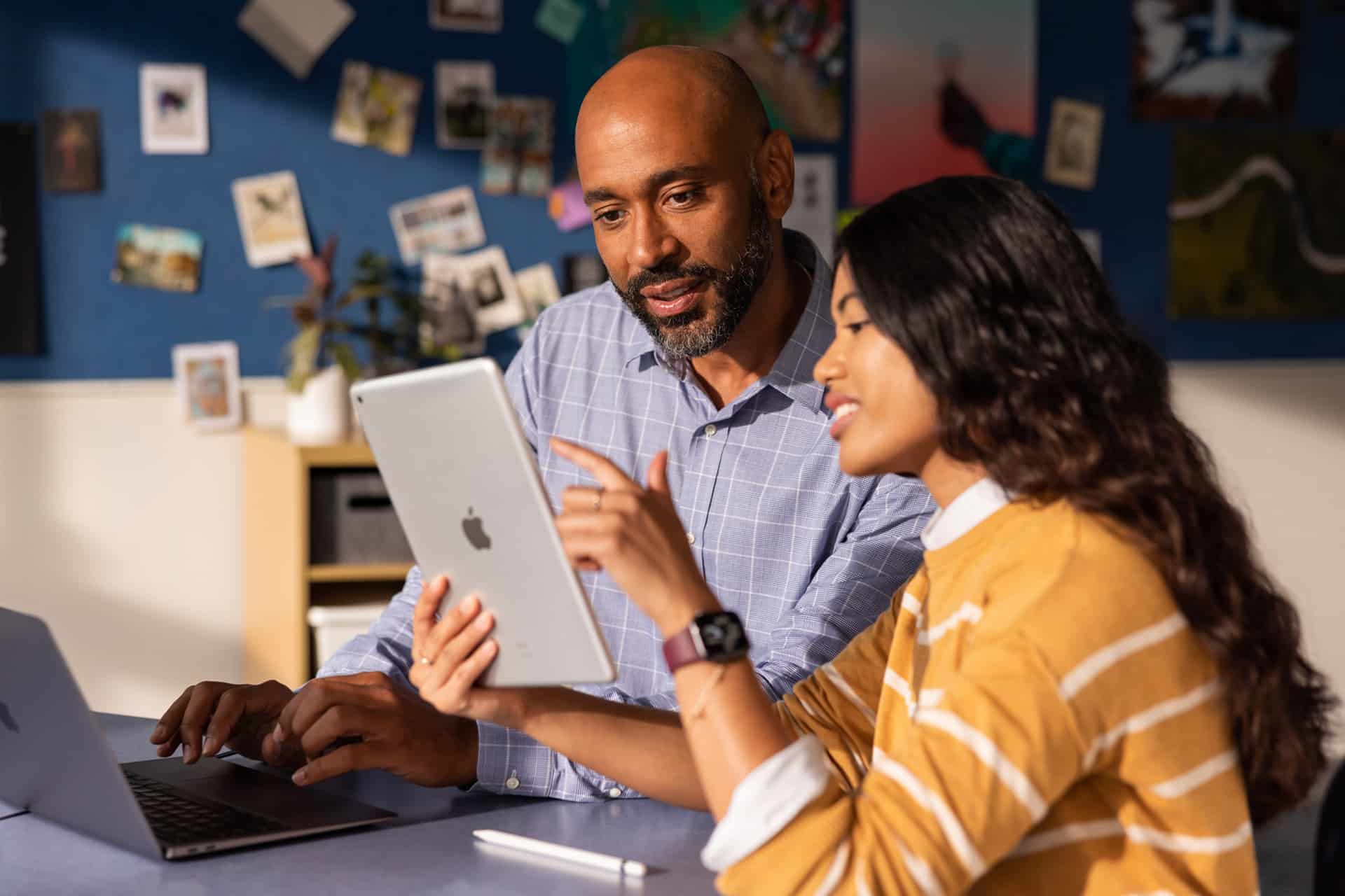 Two people sit at a table, looking at an iPad together, discussing device management strategies. One person points at the screen while the other types on a laptop, possibly configuring MDM settings for enterprise IT needs. Photos and notes are pinned on the wall behind them.